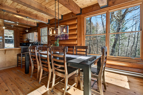 Sunlit log cabin dining for 8 with forest views and warm wood beams.