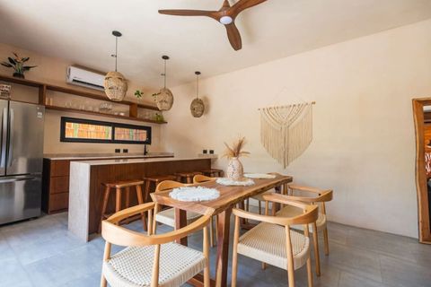 Elegant dining space with a solid wood table, woven chairs, and boho-chic decor. Rattan pendant lights and an open kitchen create a cozy, stylish ambiance.