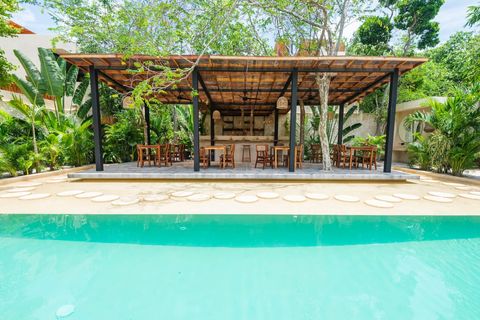 Open-air dining area with a rustic wooden pergola, surrounded by lush greenery. Overlooks a serene pool, featuring a bar and wooden seating, blending nature with comfort.