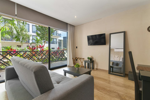 Relax in a modern living room with a cozy gray sofa, sleek coffee tables, and a flat-screen TV. Large windows fill the space with natural light and showcase lush green views.
