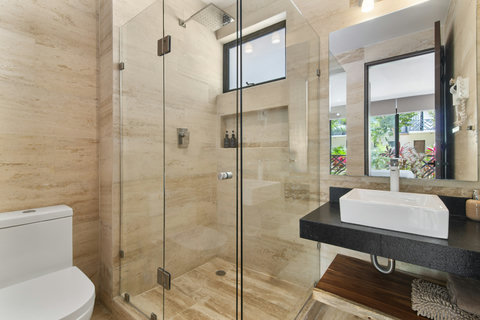 This sleek, modern bathroom features a walk-in shower with glass doors, a stylish vessel sink, and natural stone finishes. Large windows offer natural light and a view of lush greenery outside.