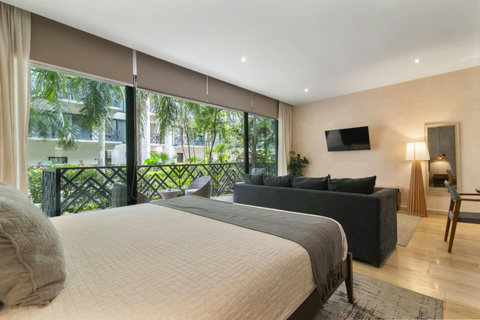 Bright and spacious studio with a king bed, lounge area, smart TV, and floor-to-ceiling windows overlooking a tropical garden and private terrace seating.