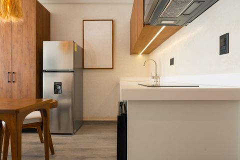 Modern kitchen with sleek white countertops, stainless steel fridge, wood cabinets, cooktop, and soft under-cabinet lighting. Includes a cozy dining area with wooden chairs.