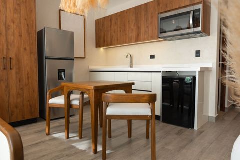 Compact kitchen with sleek white counters, wood cabinetry, stainless steel fridge, microwave, and a cozy dining table with two wooden chairs—perfect for cooking and relaxing.