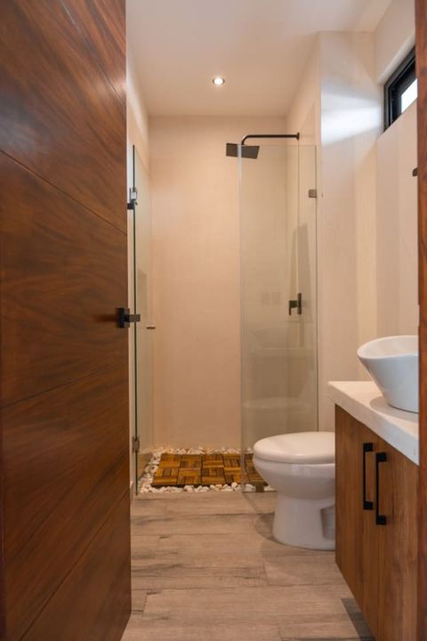 Modern bathroom with a walk-in glass shower, wood tile flooring, and white stone accents. Features a vessel sink, toilet, and warm wood cabinetry.