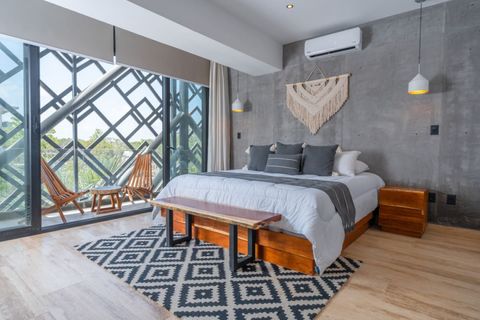 Stylish bedroom with a king bed, A/C, nightstands, pendant lighting, a bench, and private balcony. Large windows fill the room with light and offer views through a striking geometric frame.