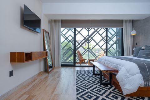 Cozy bedroom with a king-size bed, smart TV, full-length mirror, wooden bench, and balcony with chairs. Natural light fills the room through large glass doors.