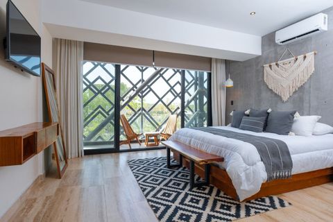 Modern bedroom with king bed, AC, flat-screen TV, balcony seating, and stylish decor featuring wood furniture and a boho macramé wall piece.