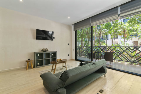 Bright living room with smart TV, stylish sofa, natural wood accents, and floor-to-ceiling windows leading to a lush private terrace—perfect for relaxing in tropical Tulum.