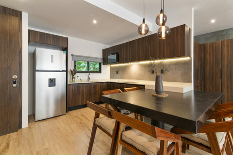 Sleek kitchen with wooden cabinets, stainless steel fridge, and a black dining table. Pendant lights and natural light create a warm, modern vibe.