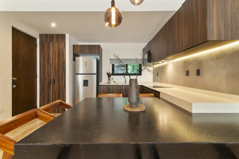 Enjoy a stylish  dining area with a sleek dining table, wooden chairs, and modern pendant lights. The open kitchen features wooden cabinets and a refrigerator, with natural light from a window.