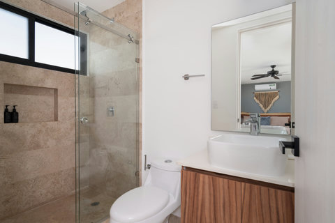 Elegant third bathroom featuring a glass shower, warm beige tiles, and natural light that enhances its airy ambiance. A bedroom view and ceiling fan complete the space with a refreshing, spa-like touch.