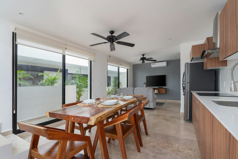 Host in style with this elegant dining area featuring a wooden table for 6, modern chairs, ceiling fans, and natural light, enhanced by lush plants that bring a fresh, nature-inspired atmosphere.