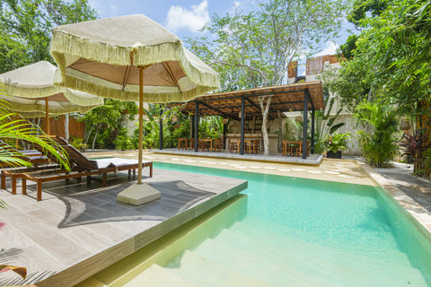 Enjoy an open-air dining area under a rustic wooden pergola, surrounded by lush greenery. Overlooking a tranquil pool, with a bar and wooden seating, it perfectly blends nature and comfort.
