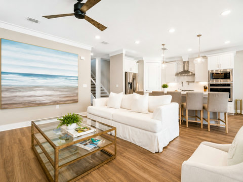 Living Room – Open-concept great room with a coastal ocean mural, plush white sofa, and gold-framed glass coffee table flowing into the kitchen.