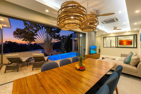 Dine under sculptural pendant lights as the sun sets, with the pool glowing just beyond the glass.