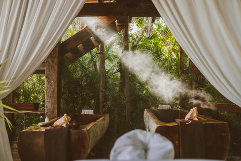 Steam drifts between carved wooden tubs as sheer curtains billow in the warm jungle air.