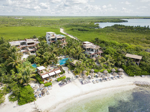 HOTEL VIEWS - Coastal retreat extends toward endless lagoon, minimalist roofs peeking through palms, shoreline carved in soft ivory curves.