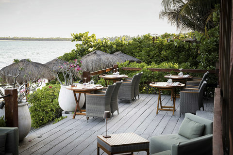 Sunlight dances across the wooden deck, where neatly set tables await amidst vibrant greenery and ocean breezes.