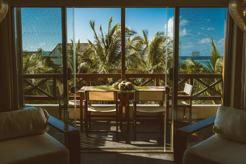 Sit at the glass-topped table beneath swaying fronds, where coconuts rest like offerings on wood grain and sunlight stripes the floor in warm, rhythmic patterns of ease.
