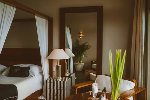 Allegra Deluxe Suite - Pause before the full-length mirror reflecting potted palms and soft-lit corners, letting the room’s layered textures, striped nightstands, trailing greens, draped muslin- wrap you in gentle stillness.