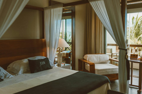 Allegra Deluxe Suite - Rest beneath a canopy where embroidered pillows meet warm wood tones, and side tables glow with metallic lamps that catch the last amber rays slipping through open terrace frames.