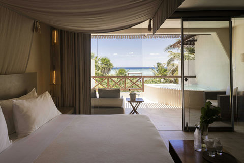 Rest beneath draped fabric that catches the breeze, while floor-to-ceiling glass dissolves boundaries between bed and balcony inviting sea air to mingle with crisp linens and slow morning stillness.