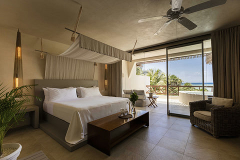 SUITE - Drift beneath suspended linen as ceiling fans stir warm air, glass doors framing ocean views while wicker armchairs and dark wood tables anchor the room in grounded, breezy ease.