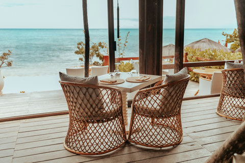PANDANO RESTURENT - Sit face-to-face at a seaside table set for two, rattan chairs cradling you as turquoise waves lap just beyond the deck — wine glasses waiting, breeze carrying salt and silence.