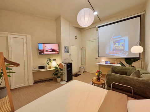 Cozy corner featuring a queen bed with fresh linens, a wardrobe for storage, and a TV. A black water purifier sits nearby, providing unlimited drinking water. The green sofa and projector screen enhance the comfort and entertainment options for group