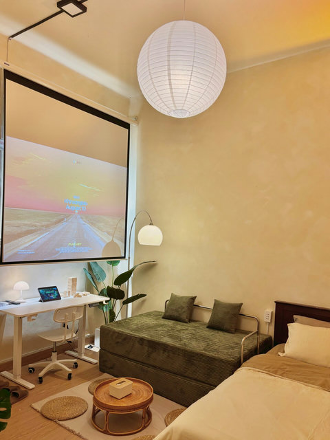 Movie nights made easy with a large projector screen right by the bed and sofa. A work desk with lamp and chair keeps things practical, while plants, soft lighting, and creamy tones add warmth, perfect for both productivity and relaxation