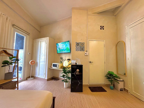 Studio corner with black water purifier and glasses, a tall white wardrobe for clothes, portable fan, and microwave on a wall shelf. Wall-mounted TV, full-length mirror, and indoor plants add function and style to the bright, neutral space.