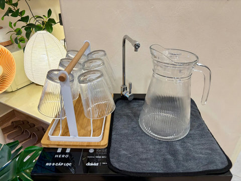 Close-up of the private water purifier with faucet, large glass jug, and a wooden rack holding six glasses. A paper lantern lamp and indoor plant add a cozy touch, offering guests unlimited safe drinking water without bottled waste