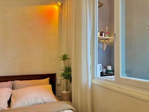 A cozy corner with a soft bed and 4 pillows, neutral bedding, and warm cream wall lighting. A tall indoor plant stands beside sheer curtains, which open to reveal a kitchenette view through the window