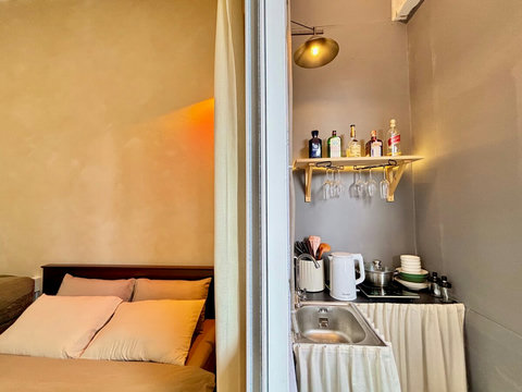 Smart layout with a queen bed next to a compact kitchenette. Features include sink, kettle, cookware, dishes, and hanging glasses under a shelf with spirits. A warm bedside glow meets practical cooking space, balancing comfort and function