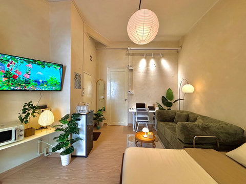 Bright studio with queen bed, green sofa, wall-mounted TV, and smart desk with chairs. Indoor plants bring freshness, while a paper lantern ceiling light and floor lamp create warm, cozy lighting for relaxation or work.