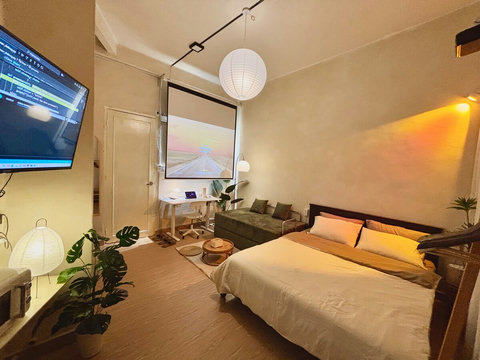 Living room with queen bed, green sofa, 4 people can sleep, and 100-inch projector screen above a smart desk. A wall-mounted TV, indoor plants, and warm lighting from ceiling and wall lamps create a cozy space for work, entertainment, and relaxation