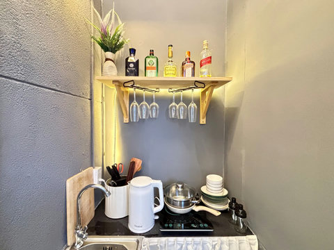 Compact kitchenette with sink, electric kettle, stovetop, and cooking utensils. Shelf above holds glasses and bottles, with plates and bowls neatly stacked. Warm lighting and a small plant complete the practical and stylish corner