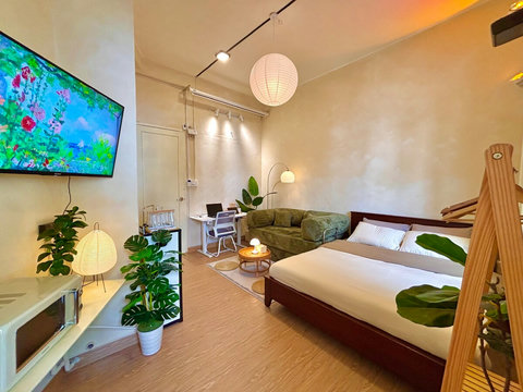 Cozy cream-toned studio with large bed in white linens, wooden rack with hangers, and a low table with four floor cushions for group dining, board games, or tea. Green sofa, plants, and soft lighting create a warm, inviting space