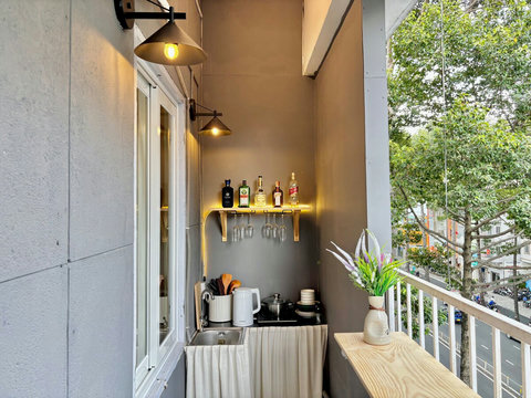 Spacious balcony with a fully furnished kitchen area, featuring a shelving for drinks and glassware. The space is illuminated with stylish lights, offering a cozy atmosphere, while lush greenery outside enhances the inviting view