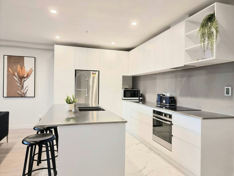 Sleek minimalist kitchen with large island bench. Built-in oven, glass cooktop, dishwasher, fridge/freezer, and microwave.