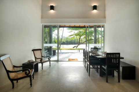 A bright dining space with serene river views. Enjoy meals at a long wooden table beside floor-to-ceiling glass doors that open to the garden and river, creating a peaceful and airy setting.