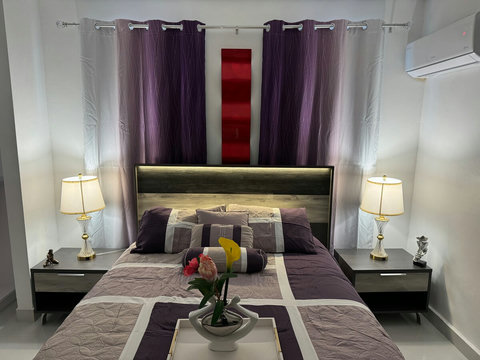 La habitación tiene una cama doble con cabecera, dos lámparas de mesa, cortinas moradas y una planta decorativa. Destaca por su iluminación cálida y esquema de color coordinado.