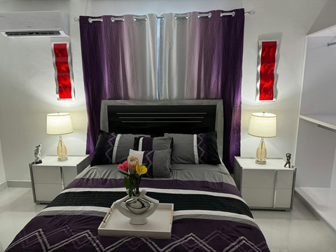 La habitación tiene una cama doble con ropa de cama morada y gris, dos mesitas de noche blancas con lámparas, cortinas moradas, un aire acondicionado y cuadros rojos. El ambiente es acogedor y bien iluminado.