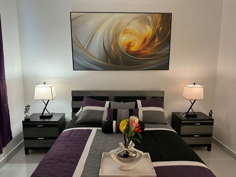 La habitación tiene una cama doble con sábanas blancas y una colcha gris. A cada lado, hay mesitas de noche con lámparas. Un cuadro abstracto grande adorna la pared sobre la cama. Una planta añade un toque natural.