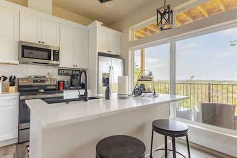The kitchen boasts modern white cabinetry, countertops, and appliances, establishing a sleek contemporary vibe. A spacious island, topped with white quartz, offers ample preparation space, accompanied by seating for informal dining.