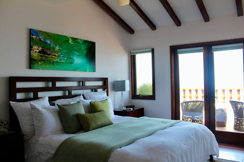 - Airy bedroom opens to a private balcony, ideal for morning coffee or sunset views
- High vaulted ceilings with exposed beams elevate the spacious feel of this serene retreat
- Features a cozy bed with layered pillows and a soothing green toned pale