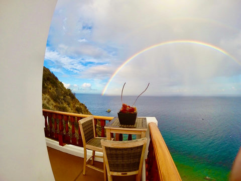 - Start your day with coffee on this serene cliffside balcony framed by a vivid rainbow and endless ocean
- Rustic wood railings and cozy chairs create the perfect nook for sea gazing
- Watch boats drift below and the sky put on a show