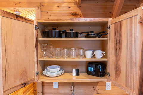 Open wooden cabinet with cookware, glassware, dishes, and a toaster in a rustic kitchen.