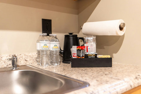 A countertop with bottled water, a black electric kettle, and a selection of coffee and tea packets next to a stainless steel sink.